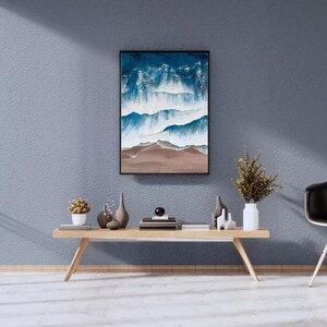 May include: A framed abstract painting of blue and white waves crashing on a sandy beach. The painting is hanging on a light blue wall above a wooden console table with decorative objects.