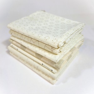May include: A stack of folded fabric squares in various off-white and beige patterns. The patterns include floral, geometric, and linear designs. The fabric appears to be cotton or a similar material, with a soft, textured appearance. The stack is neatly arranged.