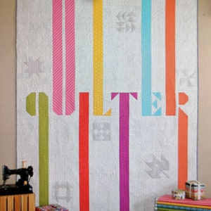 May include: A white quilt with the word "Quilter" in colorful vertical stripes. The quilt features a variety of patterns and is displayed with sewing accessories and decorative boxes. The text "Emily Herrick Designs" is at the bottom.