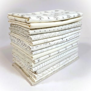 May include: A stack of folded fabric squares in various off-white and cream shades. Some fabrics feature small black patterns, including bees, crosses, and script. The fabrics are neatly stacked, showcasing a variety of textures and designs.