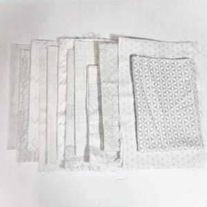 May include: A collection of white and gray fabric swatches with various patterns, including geometric shapes, lines, and floral designs. The fabrics are arranged in a layered display, showcasing the different textures and designs.
