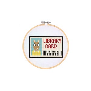 Puede incluir: Un bastidor de bordado de punto de cruz con un diseño de tarjeta de biblioteca. La tarjeta tiene un retrato de personaje de dibujos animados, las palabras "LIBRARY CARD" en rojo y un código de barras. El bastidor es de madera y tiene un fondo de tela blanca.