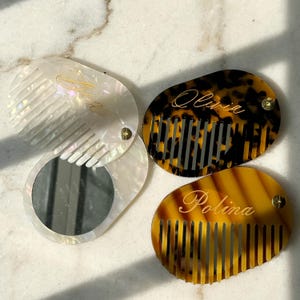 May include: Three personalised combs in different styles. One is pearlescent with a mirror, another is tortoiseshell with the name "Olivia", and the third is amber with the name "Polina". Each comb has a unique design.