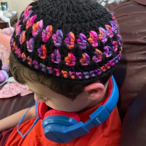 Puede incluir: Un gorro negro tejido a ganchillo con un colorido diseño de hojas en tonos rosa, naranja y morado. El sombrero lo lleva una persona que viste una camisa naranja y unos auriculares azules.