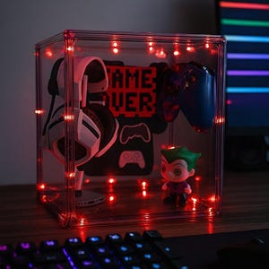Może przedstawiać: Przezroczysta gablota z czerwonymi diodami LED, prezentująca akcesoria do gier. Wewnątrz biało-czarny zestaw słuchawkowy, niebieski kontroler i figurka Jokera. Gablota zawiera napis "GAME OVER" i grafikę kontrolera.
