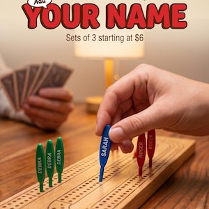 May include: A wooden cribbage board with green, blue, and red pegs. The pegs are labelled with names. The image includes the text "Personalise Your Game Night" and "YOUR NAME".