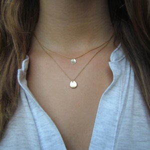 Op de afbeelding: Een gelaagde gouden ketting met twee hangercharms. De bovenste charm is een kleine cirkel en de onderste charm is een grotere, traanvormige vorm.