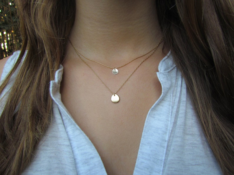 Op de afbeelding: Een gelaagde gouden ketting met twee hangercharms. De bovenste charm is een kleine cirkel en de onderste charm is een grotere, traanvormige vorm.