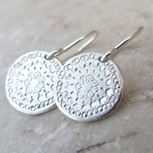 May include: A pair of silver dangle earrings with a circular design. The earrings feature a delicate floral pattern and a beaded border.