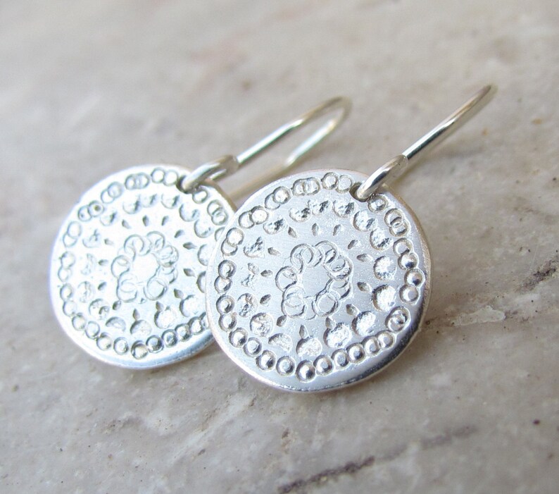 May include: A pair of silver dangle earrings with a circular design. The earrings feature a delicate floral pattern and a beaded border.