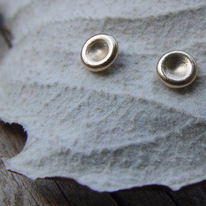 May include: A pair of gold-toned earrings with a round, slightly concave design. The earrings are displayed on a white, textured surface.