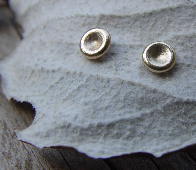 May include: A pair of gold-toned earrings with a round, slightly concave design. The earrings are displayed on a white, textured surface.