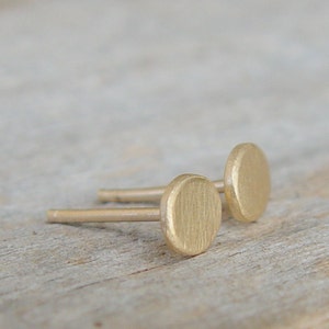 May include: A pair of gold-toned stud earrings with a round, slightly oval-shaped design. The earrings are simple and minimalist.