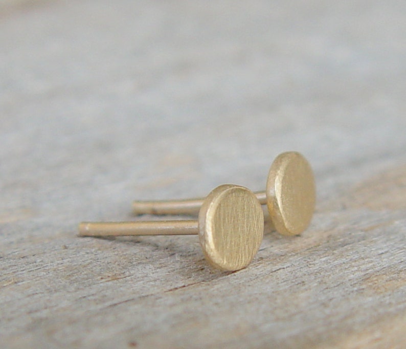 May include: A pair of gold-toned stud earrings with a round, slightly oval-shaped design. The earrings are simple and minimalist.