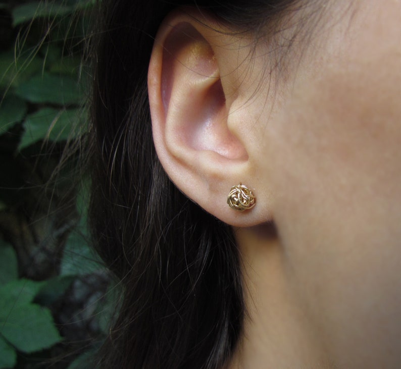 May include: A pair of gold knot-shaped earrings. The earrings are small and delicate, and they are worn in the lobes of the ears.