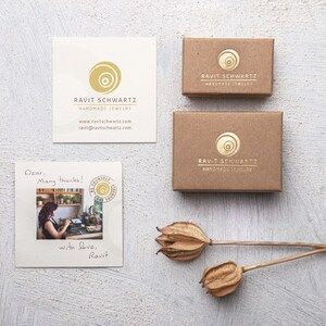 Op de afbeelding: Een bruine kartonnen doos met een gouden logo waarop "Ravit Schwartz Handmade Jewelry" staat. De doos staat op een wit oppervlak met een handgeschreven briefje waarop "Beste, Hartelijk dank! Met liefde, Ravit" staat.