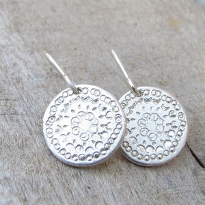 May include: A pair of silver earrings with a circular design. The earrings have a delicate, intricate pattern and a slightly textured surface.