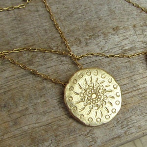 K&ouml;nnte beinhalten: Eine goldene Halskette mit einem gro&szlig;en, runden Anh&auml;nger, der ein detailliertes, kreisf&ouml;rmiges Design aufweist. Der Anh&auml;nger hat eine strukturierte Oberfl&auml;che und ein erhabenes, konzentrisches Kreisdesign.