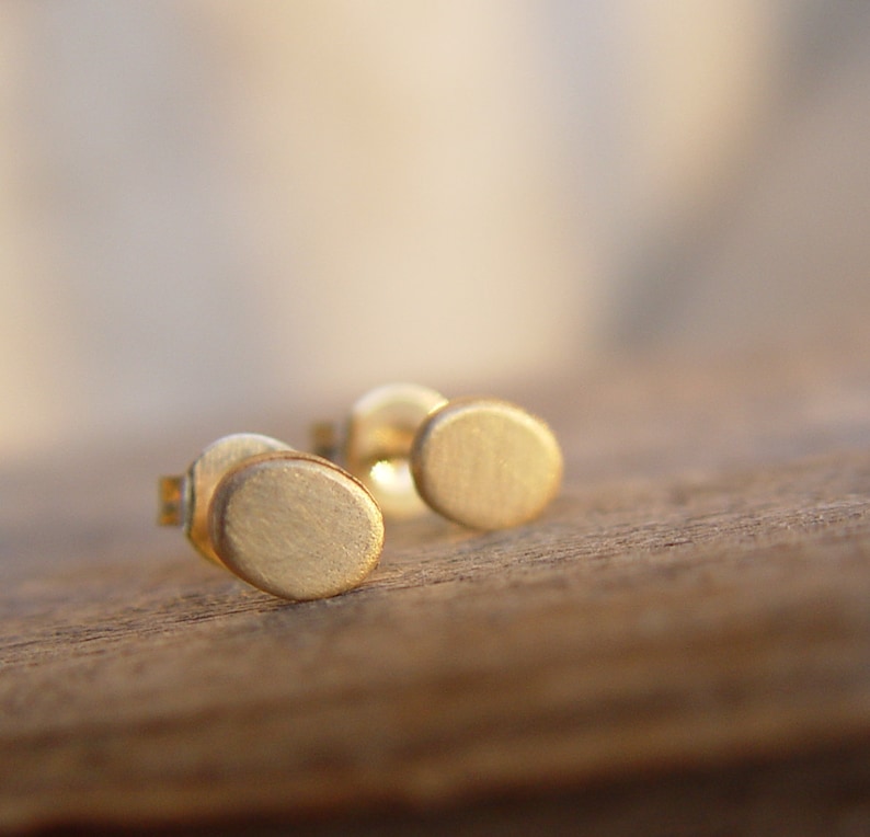 May include: A pair of gold-toned stud earrings with a circular design. The earrings are small and delicate, and they have a brushed finish.