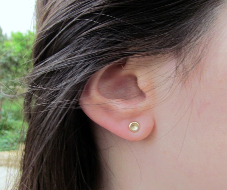 May include: A close-up of a person's ear with a small, round, gold earring. The earring has a smooth, shiny surface.