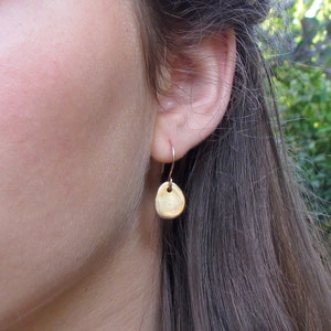 May include: Gold dangle earrings with a small, round disc pendant. The disc has a textured surface and a small hole in the center.