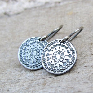 May include: A pair of silver earrings with a circular design. The earrings have a delicate wire hook and a textured, circular pendant with a raised, floral pattern.