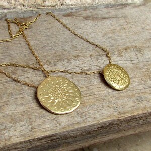 K&ouml;nnte beinhalten: Eine goldene Halskette mit zwei runden Anh&auml;ngern, die mit einem detaillierten Blumenmuster verziert sind. Die Halskette liegt auf einer Holzoberfl&auml;che.