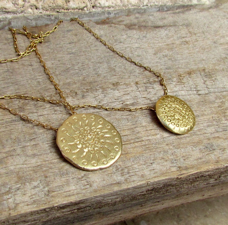 K&ouml;nnte beinhalten: Eine goldene Halskette mit zwei runden Anh&auml;ngern, die mit einem detaillierten Blumenmuster verziert sind. Die Halskette liegt auf einer Holzoberfl&auml;che.