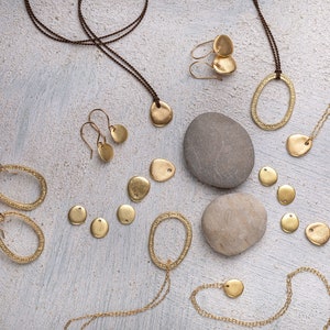 May include: A collection of gold-toned jewelry pieces, including necklaces, earrings, and pendants, arranged on a light gray textured surface with two gray stones. The jewelry features a variety of shapes, including circles, ovals, and leaves.