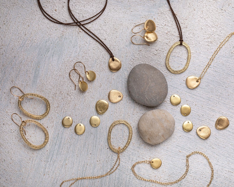 May include: A collection of gold-toned jewelry pieces, including necklaces, earrings, and pendants, arranged on a light gray textured surface with two gray stones. The jewelry features a variety of shapes, including circles, ovals, and leaves.
