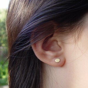 May include: A close-up of a person's ear with a small, round, gold earring. The earring is a simple, minimalist design.