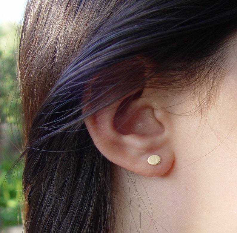 May include: A close-up of a person's ear with a small, round, gold earring. The earring is a simple, minimalist design.