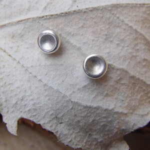 May include: A pair of silver stud earrings with a slightly rounded, concave shape. The earrings are shown on a white, textured surface.