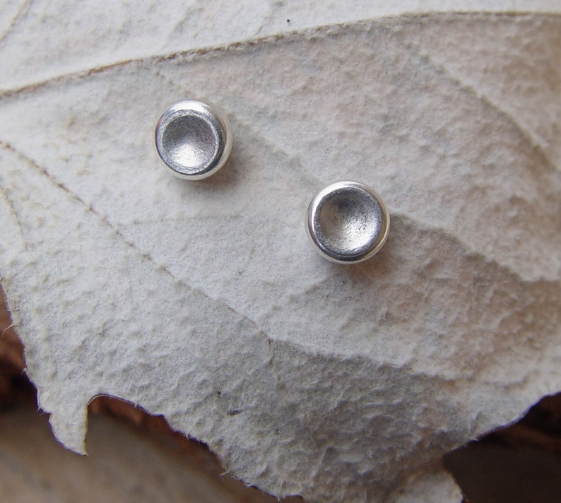 May include: A pair of silver stud earrings with a slightly rounded, concave shape. The earrings are shown on a white, textured surface.