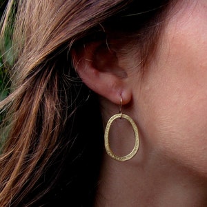 May include: Gold oval earrings with a textured surface. The earrings are hanging from a person's ear.