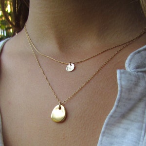 May include: Two gold necklaces with delicate chains and simple pendant charms. The larger pendant is oval shaped, while the smaller pendant is round.