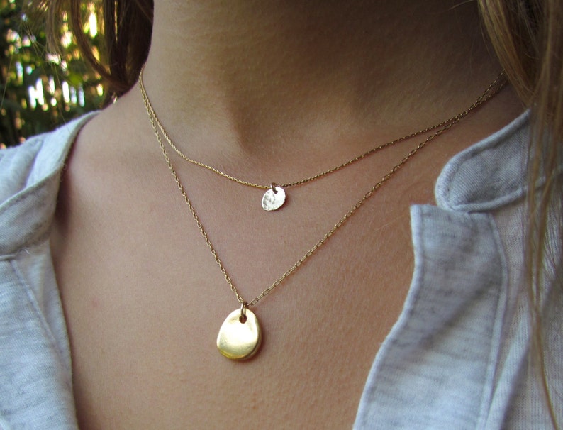 May include: Two gold necklaces with delicate chains and simple pendant charms. The larger pendant is oval shaped, while the smaller pendant is round.