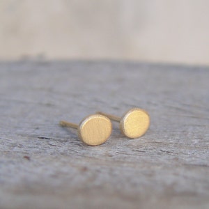 May include: A pair of small, round, gold-colored earrings. The earrings are made of a brushed metal and have a simple, minimalist design.