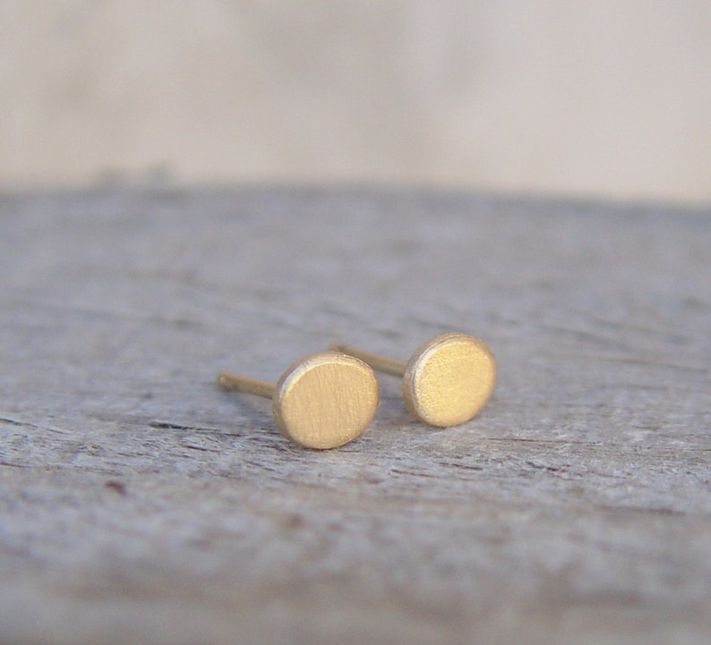 May include: A pair of small, round, gold-colored earrings. The earrings are made of a brushed metal and have a simple, minimalist design.