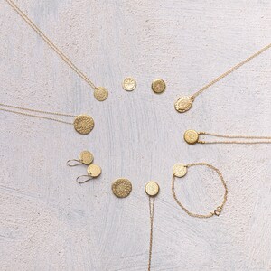 May include: A set of gold necklaces and earrings featuring circular pendants with intricate designs. The jewelry is arranged on a white textured background.