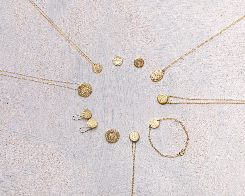 May include: A set of gold necklaces and earrings featuring circular pendants with intricate designs. The jewelry is arranged on a white textured background.