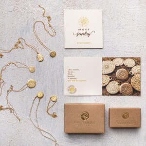 May include: A set of gold necklaces with circular pendants featuring intricate designs. The necklaces are displayed on a white textured surface with a brown card that reads "MANDALA jewelry by KIRSTI SCHMIDT" and a smaller card with a quote about mandalas. Two brown boxes with the text "KIRSTI SCHMIDT" are also shown.