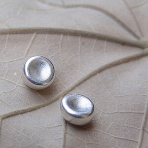 Op de afbeelding: Twee ronde, concave zilveren oorbellen met een gladde, gepolijste afwerking. De oorbellen worden getoond op een lichtbruin, getextureerd oppervlak.