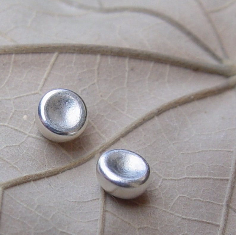 Op de afbeelding: Twee ronde, concave zilveren oorbellen met een gladde, gepolijste afwerking. De oorbellen worden getoond op een lichtbruin, getextureerd oppervlak.