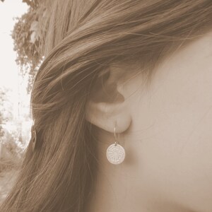 May include: A close-up of a person's ear with a gold earring featuring a round, textured disc pendant.