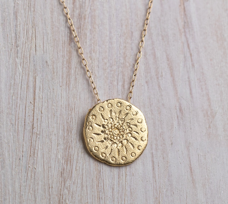 K&ouml;nnte beinhalten: Eine goldene Halskette mit einem runden Anh&auml;nger, der ein detailliertes Mandala-Design zeigt. Der Anh&auml;nger hat eine strukturierte Oberfl&auml;che und ein kreisf&ouml;rmiges Muster.