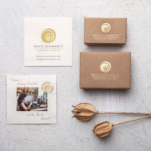 May include: A brown cardboard box with a gold logo that says "Ravit Schwartz Handmade Jewelry". The box is sitting on a white surface with a handwritten note that says "Dear, Many thanks! With love, Ravit".