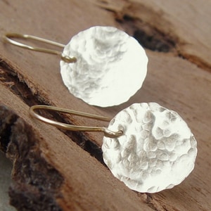 May include: A pair of silver earrings with a hammered texture and gold hooks. The earrings are round and have a slightly irregular shape.