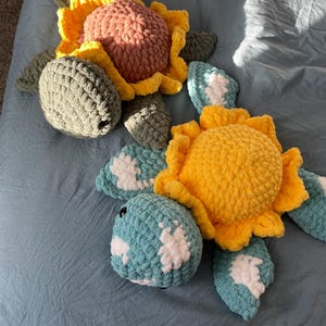 Può includere: Due peluche di tartaruga all'uncinetto con disegni di girasole. Una tartaruga è verde con un girasole rosa e giallo, e l'altra è blu con un girasole giallo.