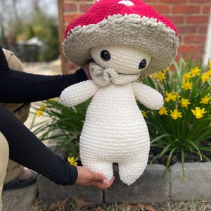 May include: A handmade crochet mushroom plushie with a red cap and white spots, a beige stem, and a small bow. The mushroom has large black eyes and is being held in front of a background of yellow flowers.